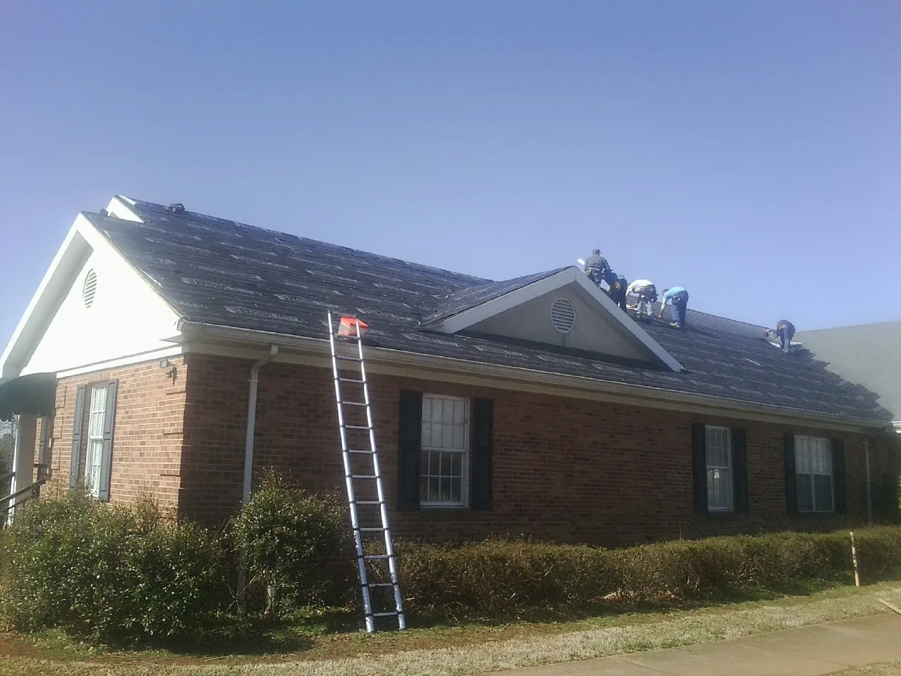 Crew tearing shingles off a brick ranch roof with a ladder set — Emergency Roof Repair work by our crew in Chatfield