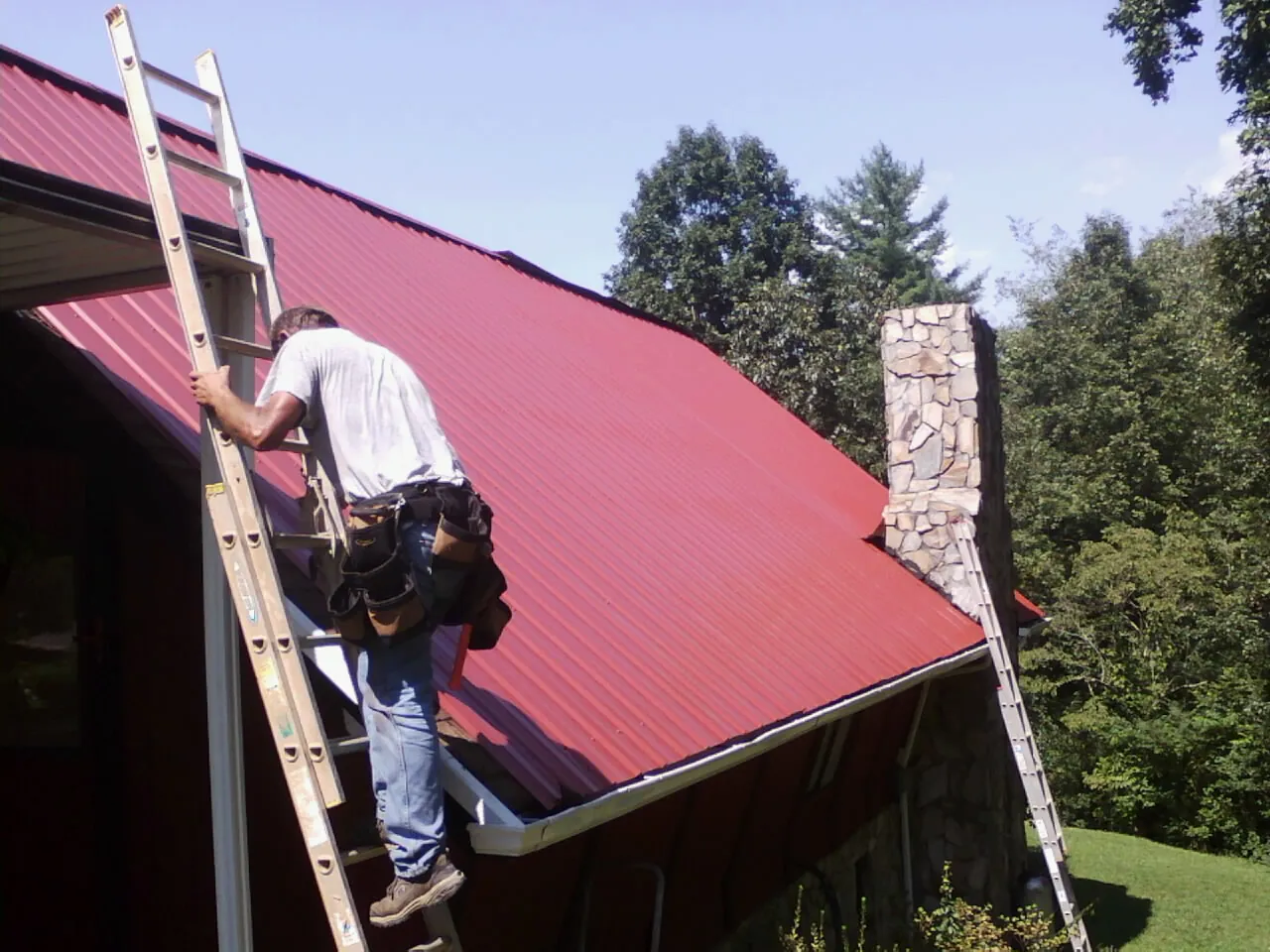 Roofer on a ladder beside a red metal roof and stone chimney — Soffit & Fascia Repair by Roofing Brothers in Chatfield